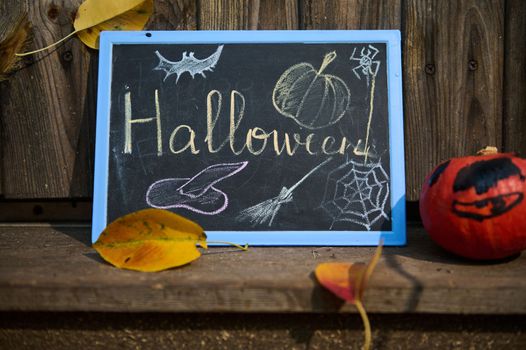 A black chalkboard, with inscription Halloween, a bright orange pumpkin and fallen dry colorful autumnal leaves on the doorstep, against a wooden background. Autumn. Halloween. Thanksgiving concept