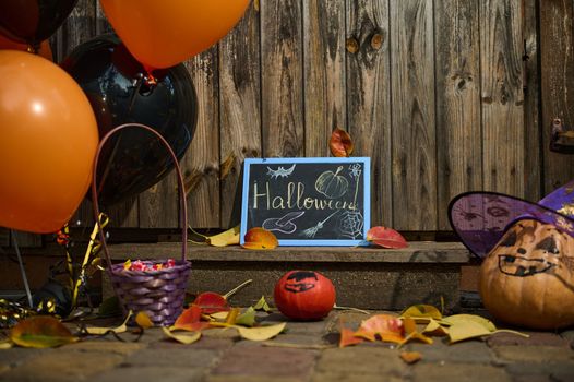 Halloween party doorstep decoration with orange black balloons, chalkboard, purple basket with candies, pumpkin with painted face, in wizard's hat and fallen autumn leaves. Halloween trick and treat