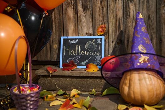 A wooden threshold or house porch, decorated for Halloween party with orange black air balloons, whole pumpkin in purple wizard's hat, fallen dry colorful autumn leaves and a violet basket with sweets