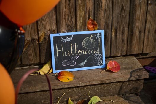 Close-up of a blackboard with chalk lettering Halloween, a pumpkin on the wooden threshold with dry autumn fallen leaves. Still life. Halloween trick and treat