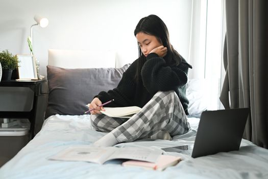 Teenage asian girl sitting on bed and doing homework. Distance education, studying online concept.