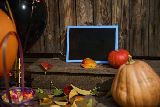 Selective focus. A blackboard with copy advertising space, next to pumpkins on a wooden threshold with fallen yellow and red leaves in autumn. Porch or backyard decoration. Halloween trick and treat