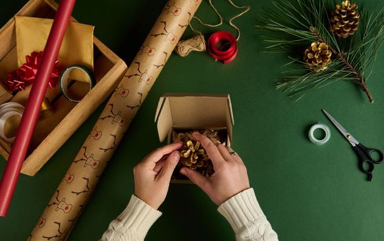 Top view woman's hands put a pine cone painted in gold, in a cardboard box while wrapping Christmas presents. Packing diy presents. New Year preparations. Celebration winter event. Handwork art craft