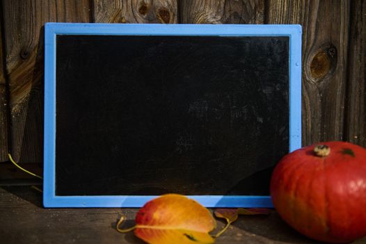 Empty black board for chalk inscriptions, with copy ad space for text, a bright orange pumpkin and fallen leaves on the threshold against a wooden background. Autumn. Halloween. Thanksgiving concept