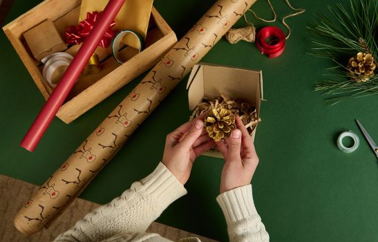 Top view woman's hands putting golden pine cone into a craft cardboard box while wrapping Christmas gifts. Packing diy presents. New Year preparations. Celebration winter event. Handwork art craft