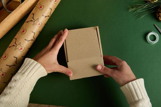 Top view Woman's hands packing Christmas or New Year gift in cardboard box, over green background with laid out wrapping decorative paper with deer pattern. Happy holiday present, surprise. Creativity