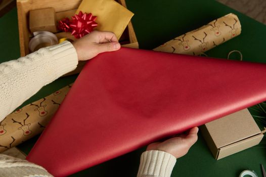 Top view woman's hands rolling out red gift paper over green surface with laid out wrapping decorative materials, bows, tapes and ribbons. Diy presents, Handwork art. New Years, Christmas preparations