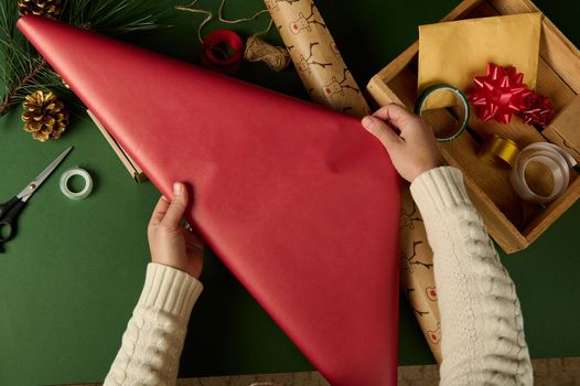 Top view woman's hand rolling out a red gift paper while, choosing wrapping decorative materials, over green background. Golden envelope, tied bow and tapes on wooden crate. Packing Xmas diy presents