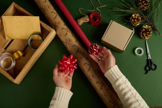 Top view woman's hands holding tied bows over wrapping paper with deer pattern and of red color, on a green surface with materials for packing Christmas and New Year gifts. Boxing Day. Handwork. Diy