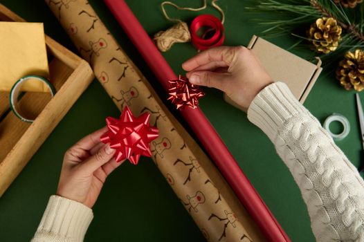 Top view woman's hands apply tied bows on wrapping paper with deer pattern and of red color, on a green surface with materials for packing Christmas and New Year gifts. Boxing Day. Handwork art. Diy