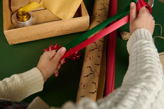 Top view of woman's hands, holding red and green satin decorative tapes over wrapping gift paper, packing presents for Christmas or New Year. Diy. Creative idea. Handwork art. Boxing Day. Creativity