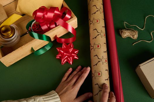 Close-up woman's hands choosing wrapping paper with deer pattern and of red color, next to a wooden crate with decorative tapes, ribbons and bows on a green background. Packing presents. Christmas