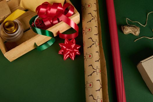 Close-up wrapping paper with deer pattern and of red color, next to a wooden crate with decorative tapes, ribbons and bows on a green background. Boxing Day. Packing presents. Christmas magic mood