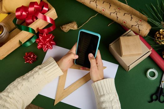 Selective focus on woman's hands holding a smartphone empty blank touch screen, with copy ad space for promotional text, over a green background with Xmas wrapping materials, decorative tapes and bows