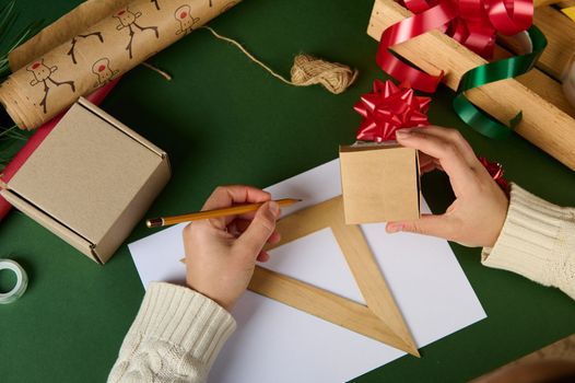 Top view woman's hands, holds cardboard box, calculates the amount of wrapping material for packing gifts for Christmas, Women's, Saint Valentine's Day or any other event using wooden ruler and pencil