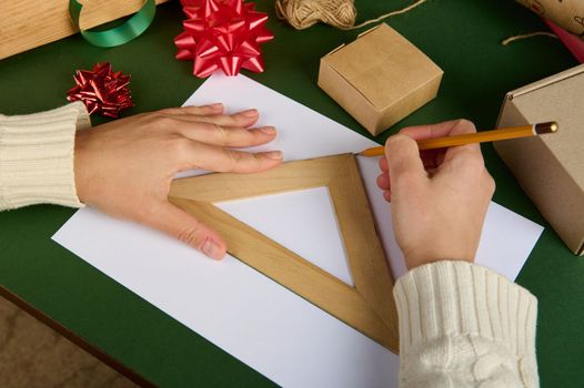 Top view woman's hands drawing on white paper using a ruler and a wooden pencil, on a green surface with wrapping material for packing Christmas, New Year gifts. Diy presents for any celebration event