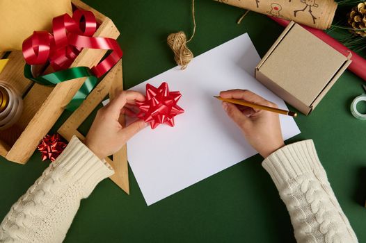 Close-up woman using wooden crayon, drawing, writing on a white paper sheet with copy ad space, on green surface, with wrapping creative and decorative materials, ornaments for packing Christmas gifts