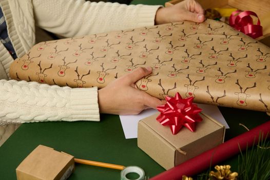 Close-up of woman's hands rolling out a wrapping paper with deer pattern while packing Christmas gift over a background with decorative materials. Boxing day. Diy presents. Celebration winter event