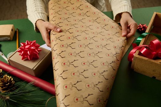 Close-up woman rolling out a wrapping gift paper with deer pattern while packing Christmas presents. Boxing Day. New Year and Xmas preparations. Happy winter holidays. Diy presents. Handmade art work