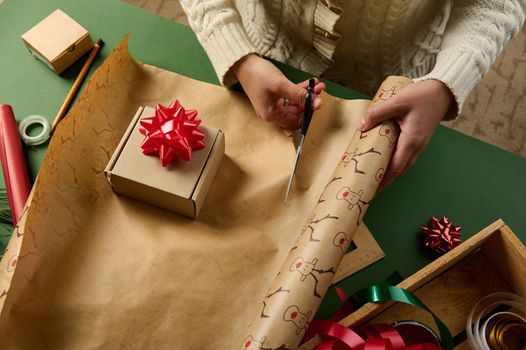 Overhead view of a woman cutting the wrapping paper while packing gifts for Christmas, New Year or any other celebration event. Decorative tapes on a wooden crate in green background. Boxing Day