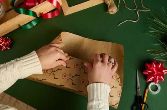 Top view of woman's hands wrapping a present for Christmas or New Year's events, using beautiful decorative paper with deer pattern, over green background. Boxing Day. Xmas preparations. Copy ad space