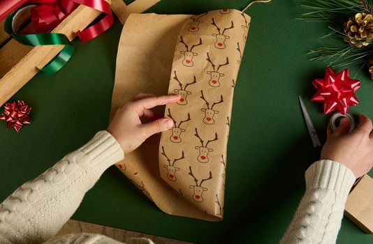 Top view of woman's hands wrapping a present for Christmas or New Year's events, using beautiful decorative paper with deer pattern, over green background. Boxing Day. Xmas preparations. Copy ad space