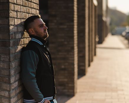 Caucasian bearded man in a bomber jacket leaned against a brick wall