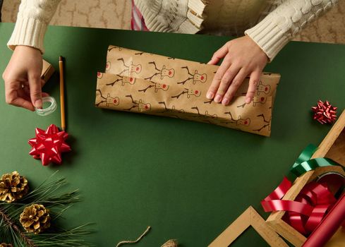 Close-up woman wrapping gift for Christmas in a paper with deer pattern, on a green background with golden pine cones as Xmas decoration, tied bow and decorative tapes in a wooden crate. Boxing Day
