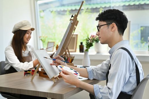 Young creative man attending in art workshop, painting picture with watercolor on canvas.