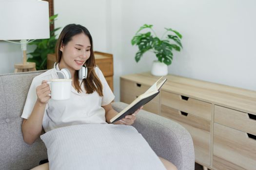 Asian woman reading book and wearing headphones and drink coffee cup relaxing on sofa at home.