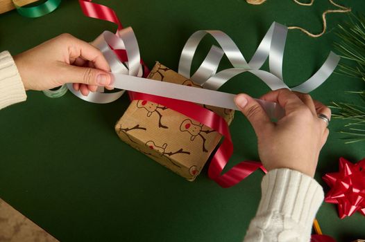 Top view of woman's hands, holding red and white satin decorative ribbons over wrapping gift paper, packing presents for Christmas or New Year. Diy. Creative idea. Handwork art. Boxing Day. Creativity