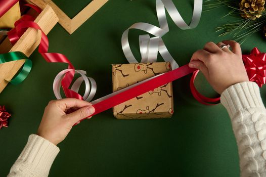 Top view woman in warm beige sweater, choosing the decorative ribbon for tying up a Christmas present, wrapped in a wrapping paper with deer pattern over green background. Boxing Day