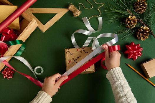 View from above of a woman in warm beige sweater, choosing the decorative ribbon for tying up a Christmas present, wrapped in a wrapping paper with deer pattern over green background. Boxing Day