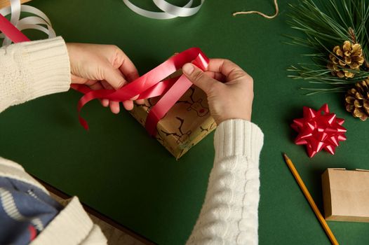 Top view woman's hands tying up a present wrapped in a paper with deer pattern, using a red shiny ribbon. Christmas and New Year preparations. Diy. Creative idea. Handwork art. Boxing Day. Creativity