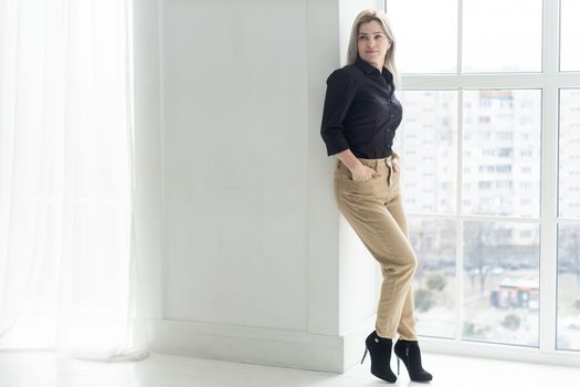 Full length view of young woman standing near window, wearing black, resting from the business.