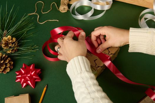 Overhead view of woman's hands tying up a christmas present with a decorative shiny red ribbon, over a green background with laid out wrapping materials. Handwork creative art. Packing diy presents