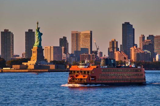 Image of Large Staten Island Ferry passing by Statue of Liberty in golden hour of New York City with skyline behind