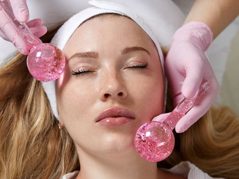 Young woman receiving facial massage with glass balls in beauty salon, concept of face massage in spa