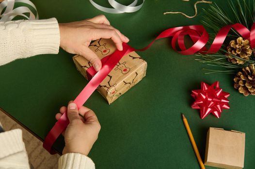 Close-up woman using a shiny red ribbon, ties up a gift wrapped in wrapping paper with deer pattern, over green background. Copy advertising space. Christmas and New Year's preparations. Boxing Day