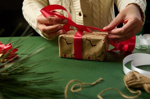 Close-up woman using a shiny red ribbon, ties up a gift wrapped in wrapping paper with deer pattern, over green background. Christmas and New Year's preparations. Boxing Day. New Year. Diy presents