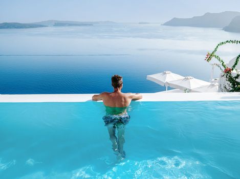 Santorini Greece Oia, young men in swim shorts relaxing in the pool looking out over the caldera of Santorini Island Greece, infinity pool, a young guy on a luxury vacation in Europe Greece. 