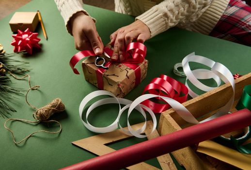 Close-up woman wraps Christmas gift box, tying up with a shiny red ribbon. Decorative tapes and bows, linen rope and ornaments on green background. New Year preparations. Packing presents. Boxing Day