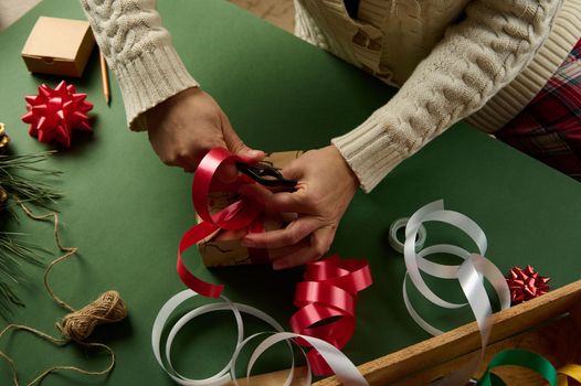 Top view woman in warm beige sweater, wrapping a Christmas or New Year's gift, tying a beautiful bow from a red shiny decorative ribbon. Boxing Day. Diy presents. Magical atmosphere of winter holidays