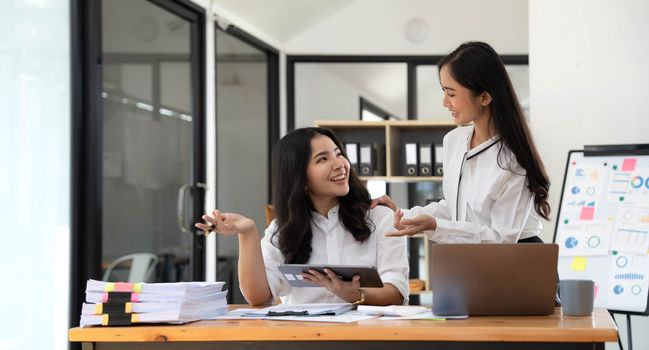 Two young Asian businesswomen show joyful expression of success at work smiling happily with a laptop computer in a modern office..