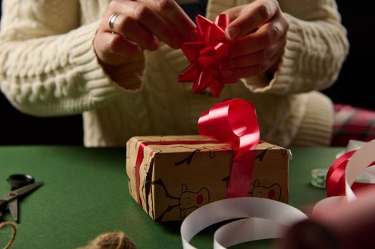 Close-up. Front view of woman's hands decorating a Christmas gift with beautiful tied bow. Packing presents for New Year or any other celebration event. December 25th. Boxing Day.