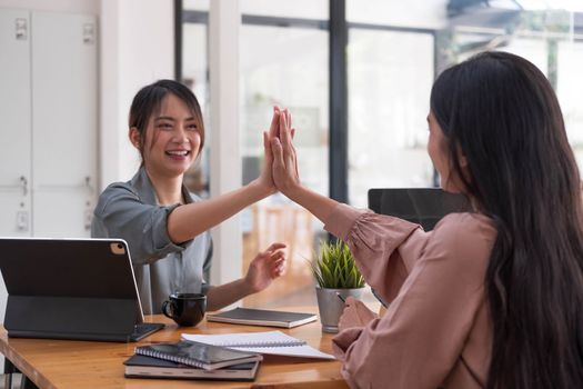 Two young Asian businesswomen show joyful expression of success at work smiling happily with a laptop computer in a modern office...