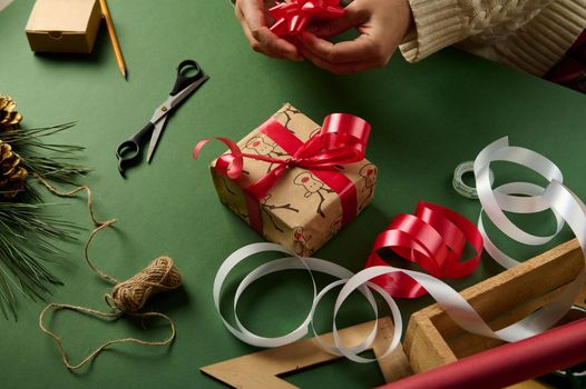 Packaging Christmas presents over a green background with decorative tapes, linen rope and wrapped material lying on a wooden crate. New Year's preparations. Winter holidays. Boxing Day