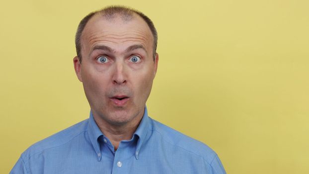 Portrait of a surprised handsome man 45-55 years old in a blue shirt looks at the camera.A man in a blue shirt on a yellow background.