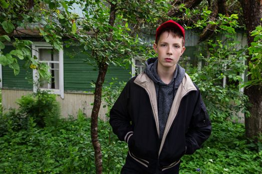 A boy in a demi-season jacket looks strictly at the camera against the backdrop of a village house. Rest of the boy in the village. boy on the background of foliage and a country house