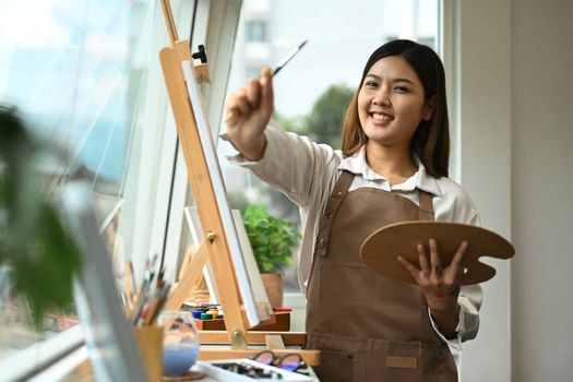 Cheerful young woman enjoying creative leisure activities, painting picture in bright modern art studio.